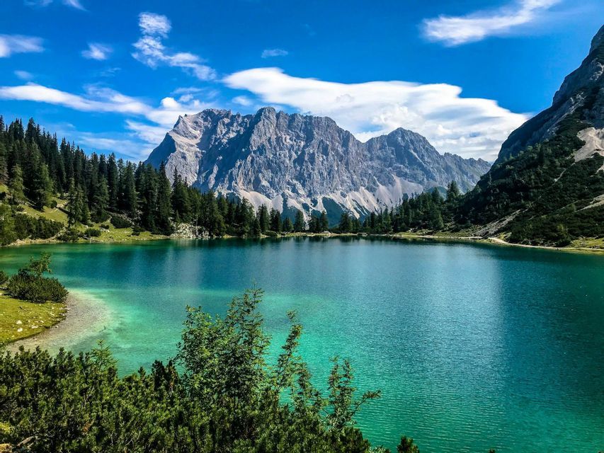 Un lago alpino turchese circondato da una pineta verde, alla base di una grande montagna rocciosa sotto un cielo azzurro.
