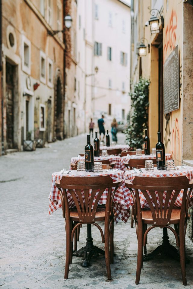 Tavoli con tovaglie a quadretti bianchi e rossi sono apparecchiati con bottiglie di vino in un caffè all'aperto su una strada acciottolata.
