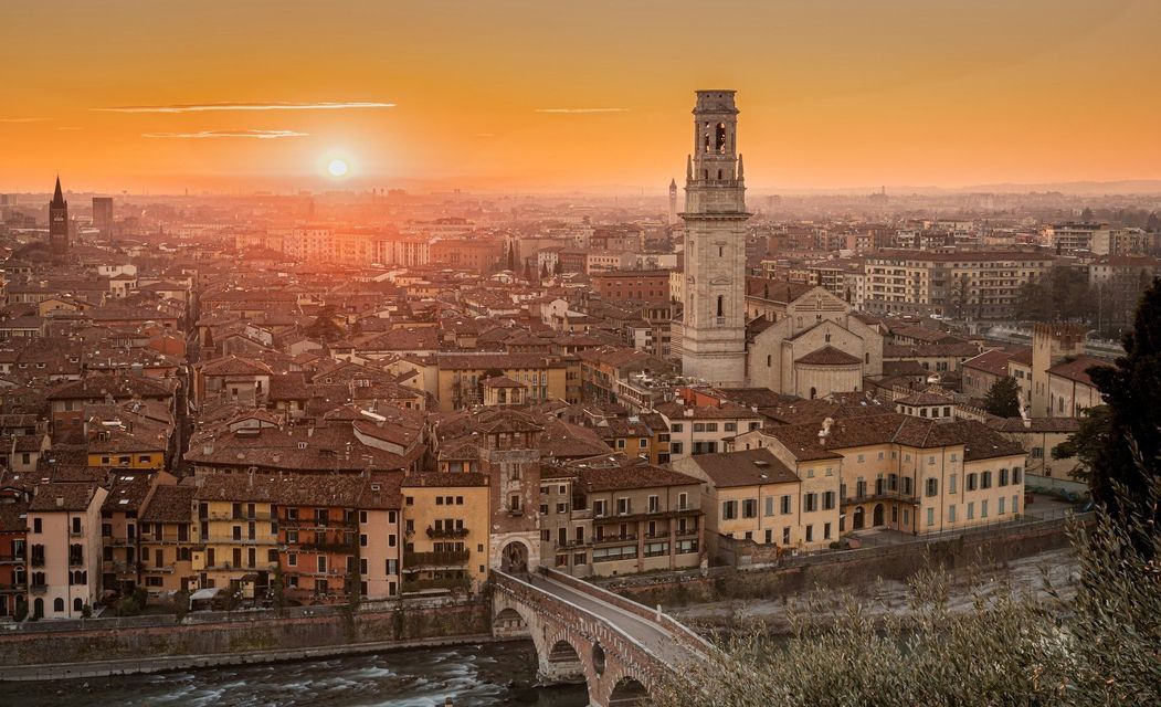 Una veduta aerea di una città storica con tetti in terracotta, un ponte di pietra su un fiume e un imponente campanile al tramonto.
