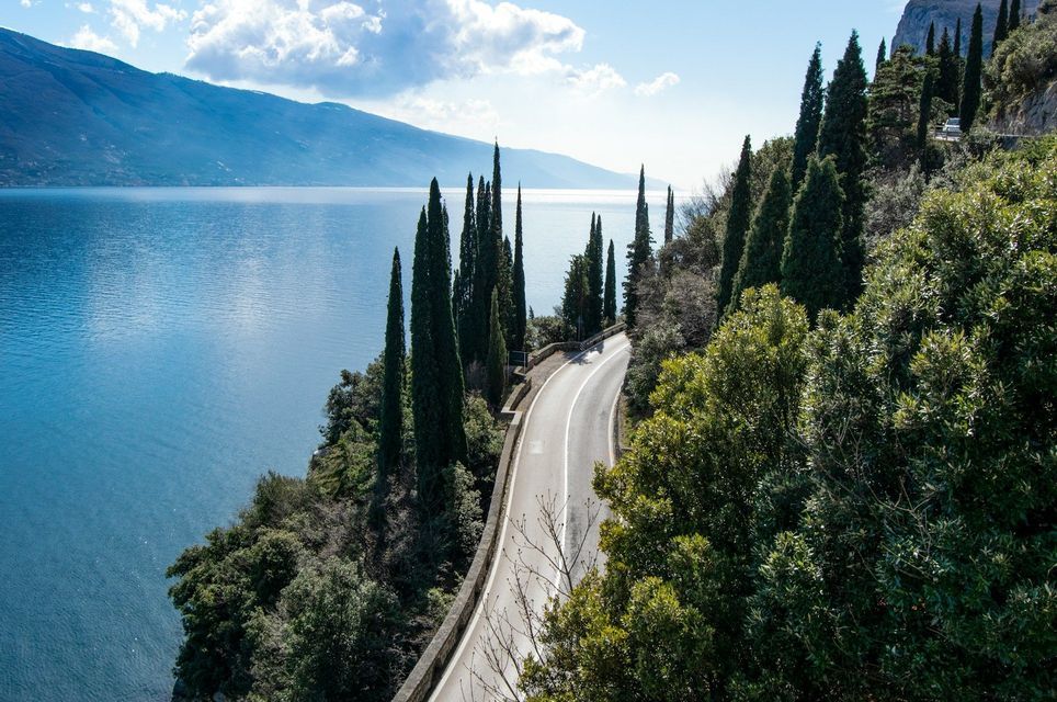 Veduta aerea di una strada tortuosa in riva al lago circondata da cipressi, con montagne visibili al di là dell'acqua.