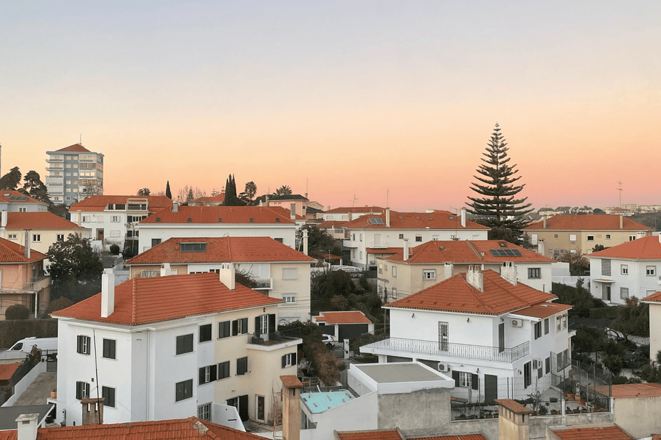 A dense neighborhood of white houses with red-tiled roofs sits on a hillside against a soft, pastel sunset sky.
