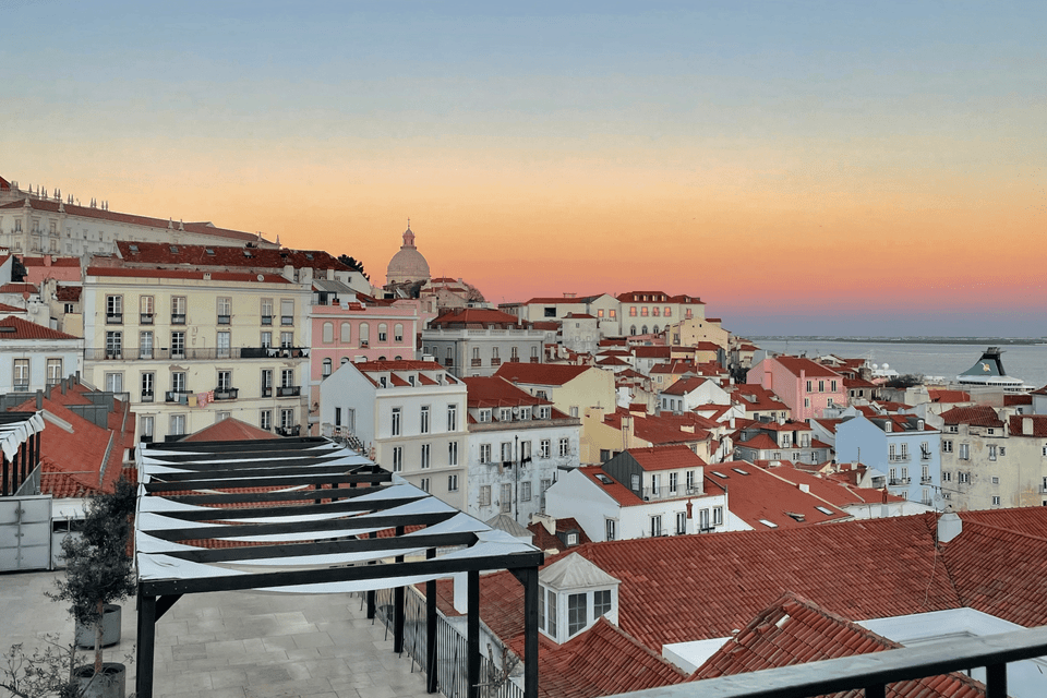 A cityscape of colorful buildings with red-tiled roofs seen from a terrace at sunset, with the sea in the distance.
