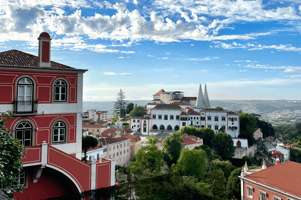 A red and white building overlooks a historic town with a palace featuring two white conical chimneys, under a partly cloudy sky.