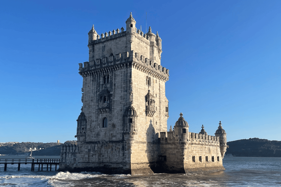 An ornate stone fortress with turrets stands in the water next to the shore under a clear blue sky.