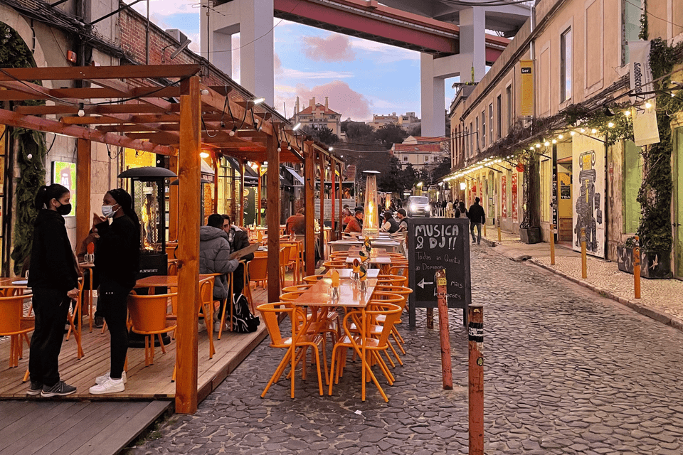 An outdoor restaurant with bright orange chairs is set up on a cobblestone street under a large bridge at dusk.