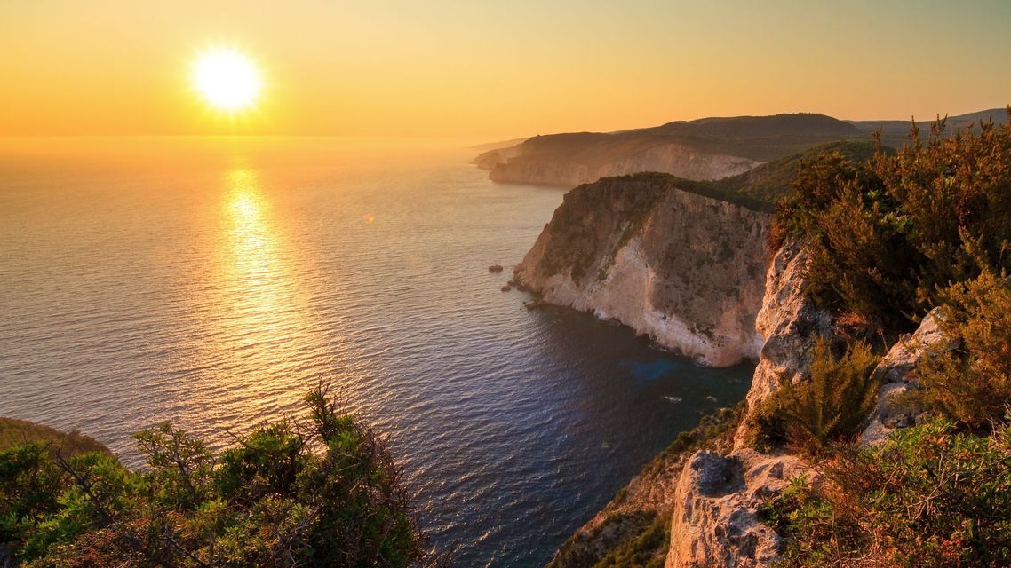 Il sole tramonta sull'oceano, con il suo riflesso che scintilla sull'acqua accanto a una costa rocciosa e ricoperta di arbusti.