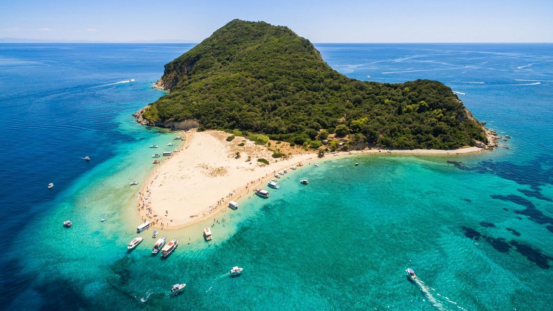 Veduta aerea di un'isola verde lussureggiante con una lingua di sabbia dove numerose barche sono ormeggiate in acque turchesi vicino alla costa.