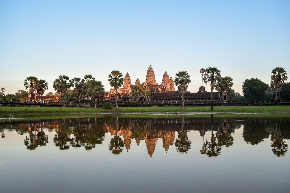 Un grand complexe de temples en pierre et des palmiers environnants se reflètent parfaitement dans un lac calme sous un ciel bleu clair.