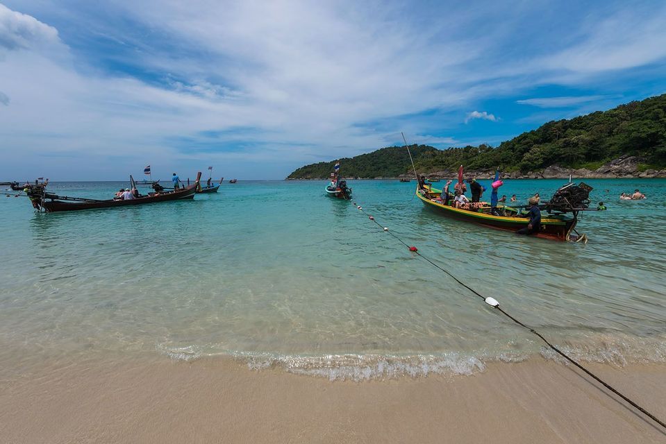 Varias barcas de cola larga con un grupo de viaje de WeRoad flotando en agua turquesa en una playa de arena con una colina boscosa detrás.