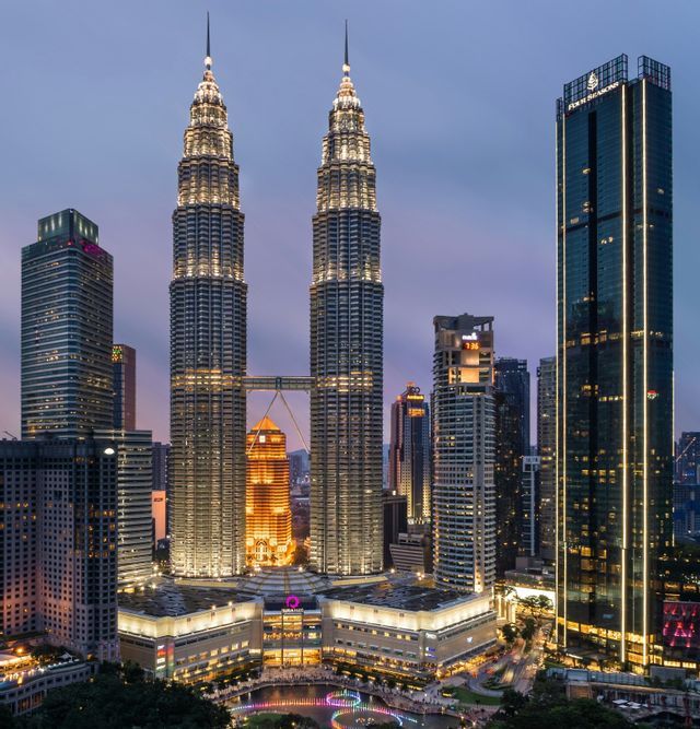 Les Tours Petronas illuminées et les gratte-ciel environnants composent une ligne d'horizon urbaine moderne au crépuscule, avec une fontaine colorée en contrebas.