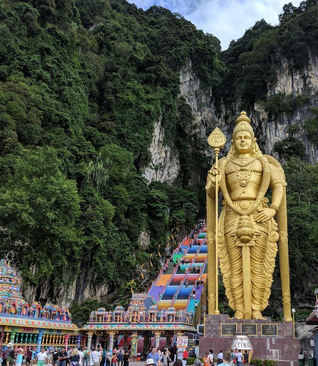Une grande statue dorée d'une divinité se dresse près d'un long escalier aux couleurs de l'arc-en-ciel, menant à une falaise boisée et escarpée.