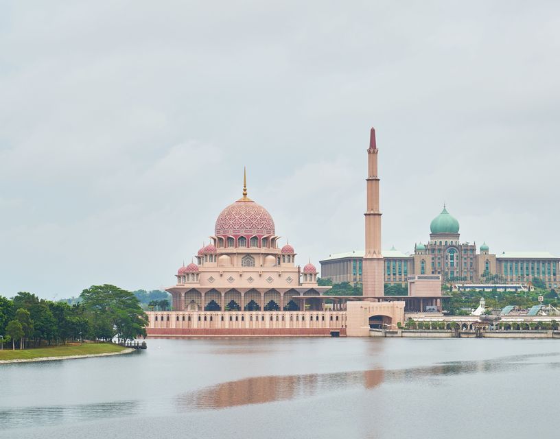 Une grande mosquée aux dômes roses ouvragés et à un haut minaret se dresse au bord d'une étendue d'eau sous un ciel nuageux.