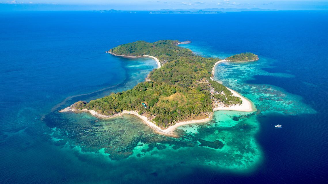 Una vista aerea di un'isola tropicale lussureggiante e verde con spiagge di sabbia bianca, circondata da acque limpide turchesi e blu intenso.