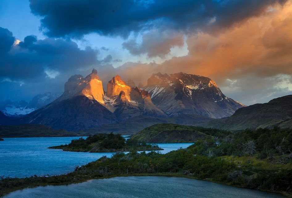 Cime montuose frastagliate illuminate dalla luce dorata del sole sotto un cielo scuro e nuvoloso, con un lago blu e calmo in primo piano.