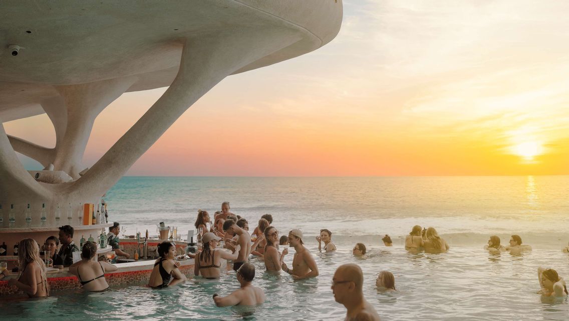 Un gruppo WeRoad che socializza a un moderno swim-up bar in una piscina a sfioro con vista sull'oceano al tramonto.