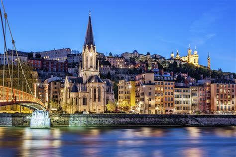 Un paesaggio urbano al tramonto con una chiesa illuminata e edifici lungo un fiume, con luci che si riflettono sull'acqua.