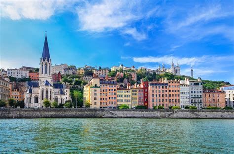 Un paesaggio urbano con edifici colorati e una chiesa con un alto campanile che costeggiano un fiume sotto un cielo azzurro.