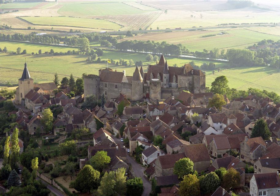 Veduta aerea di un borgo medievale con un grande castello in pietra su una collina, circondato da una verde campagna.