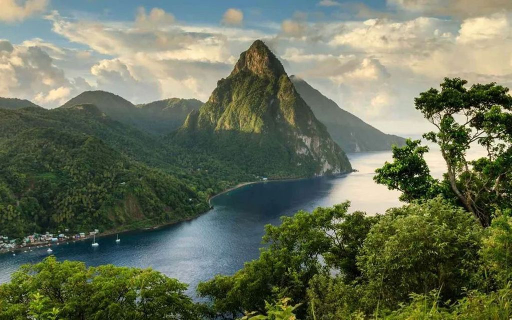 Una grande montagna conica verde sulla costa di una baia blu, con un piccolo villaggio e barche a vela sull'acqua.