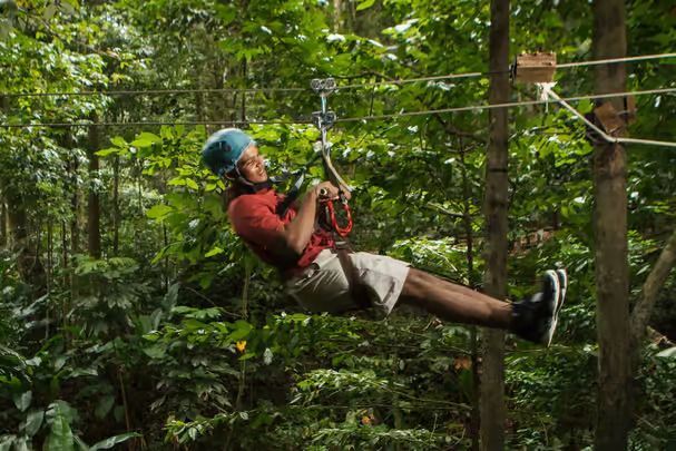 Un uomo con casco e imbracatura blu sorride mentre scende in zipline su un cavo attraverso una fitta giungla verde.