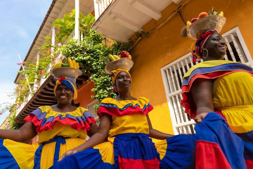 Tre donne con abiti tradizionali colorati bilanciano cesti di frutta sulla testa sorridendo in una strada di città.