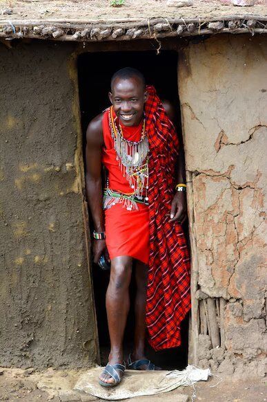 Un uomo sorridente, in abito tradizionale rosso e gioielli di perline, si trova sulla soglia di una capanna di fango.