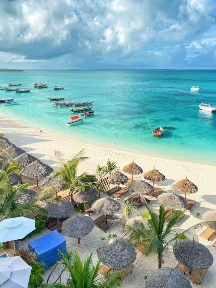 Una vista dall'alto di una spiaggia tropicale di sabbia bianca con ombrelloni di paglia e barche che galleggiano su acque calme e turchesi.
