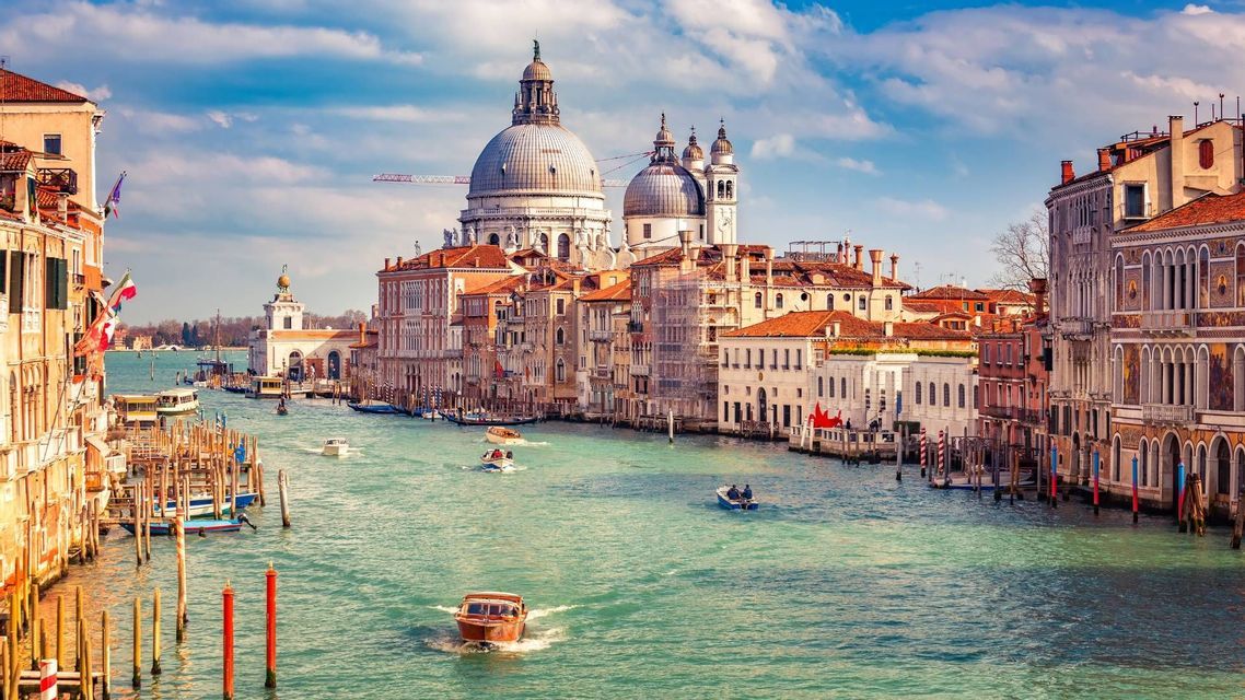 Un ampio canale con diverse barche è fiancheggiato da edifici storici, con una grande basilica a cupola sullo sfondo sotto un cielo azzurro e nuvoloso.