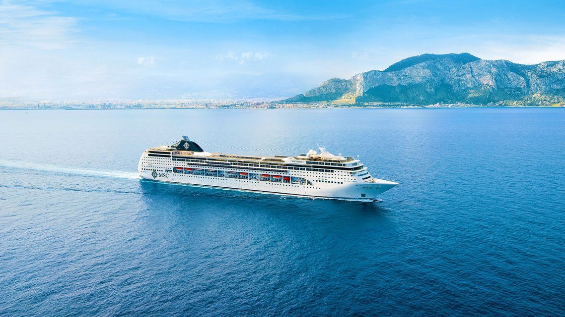 Una grande nave da crociera bianca naviga su un oceano blu calmo con una costa montuosa distante sullo sfondo.