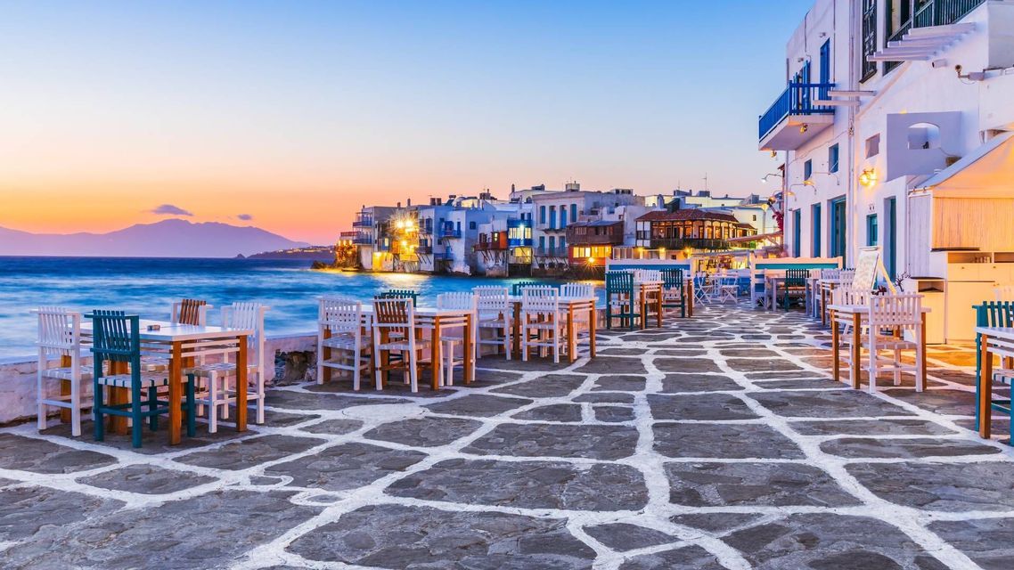 Tavoli e sedie vuoti su un patio acciottolato con vista sul mare e su una città costiera al tramonto.