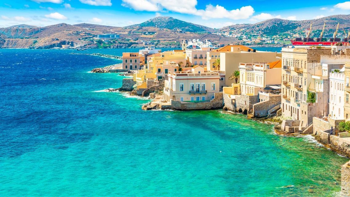 Una città costiera con edifici gialli e bianchi si trova su una costa rocciosa accanto a un mare turchese sotto un cielo blu e parzialmente nuvoloso.