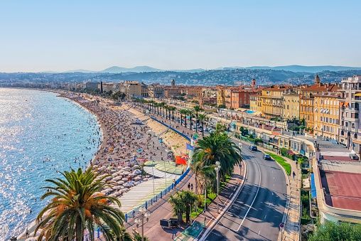 An aerial view of a crowded beach beside a sparkling blue sea, a coastal road, and a city skyline under a clear sky.