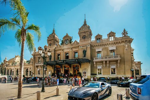 Luxury cars are parked in a plaza in front of an ornate, classical-style building under a clear blue sky.