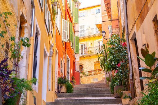 A narrow stone staircase ascends through an alleyway lined with colorful yellow and orange buildings and potted plants.