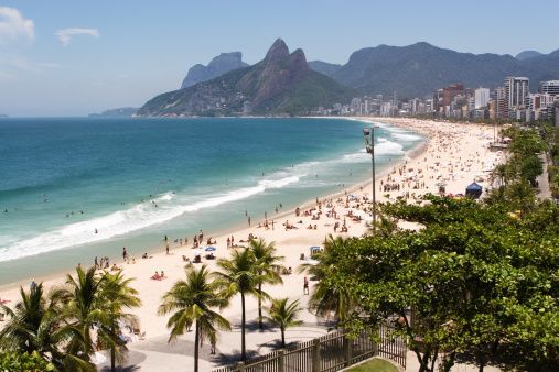 An elevated view of a crowded sandy beach with people swimming, backed by a city and large mountains under a blue sky.