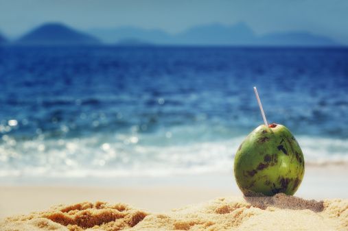 A green coconut with a straw sits on a sandy beach, overlooking the blue ocean and distant mountains.