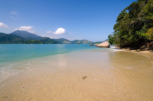 A sandy beach with clear, shallow water leading to a bay with green mountains in the distance under a blue sky.