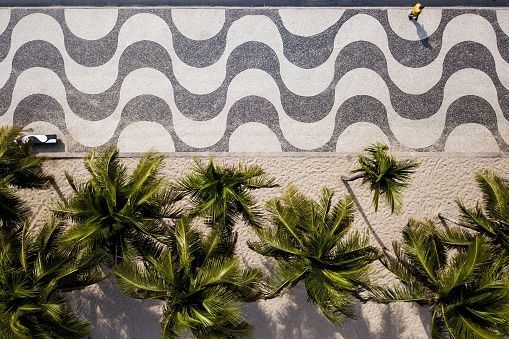 A top-down aerial view of a wavy black-and-white patterned sidewalk next to the tops of palm trees on a sandy beach.