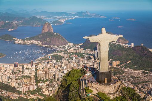 The Christ the Redeemer statue, viewed from behind, overlooks a sprawling cityscape, a bay, and Sugarloaf Mountain.
