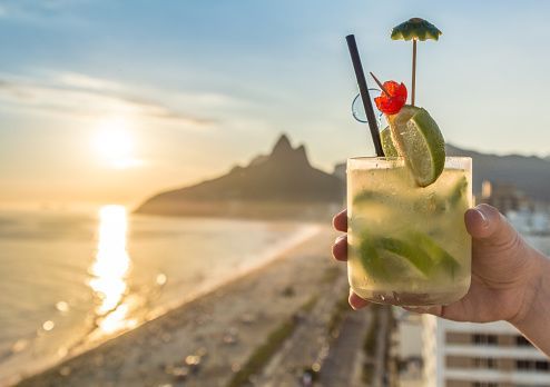 A hand holds a garnished lime cocktail overlooking a beach with mountains in the background at sunset.