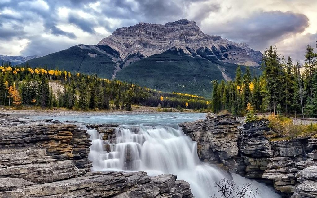 Uno scatto a lunga esposizione di una cascata che si getta su rocce stratificate, con un paesaggio montano e boscoso sotto un cielo nuvoloso.