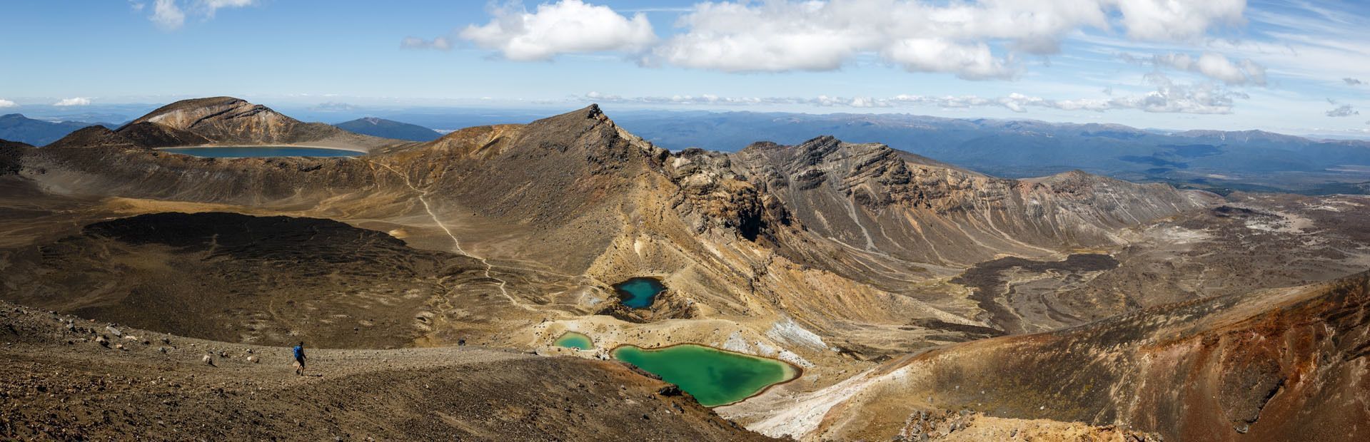 Ein Panoramablick auf eine Vulkanlandschaft mit mehreren grünen und blauen Kraterseen, mit einem Wanderer, der im Vordergrund auf einem Pfad geht.