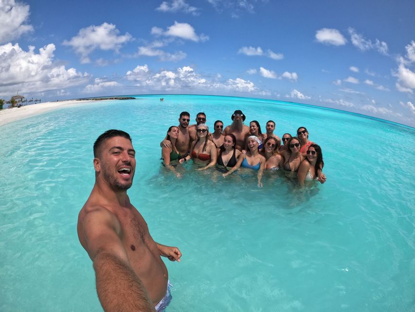 Ein Mann macht ein Selfie von einer lächelnden WeRoad-Gruppe, die gemeinsam im türkisfarbenen Meerwasser nahe einem weißen Sandstrand steht.