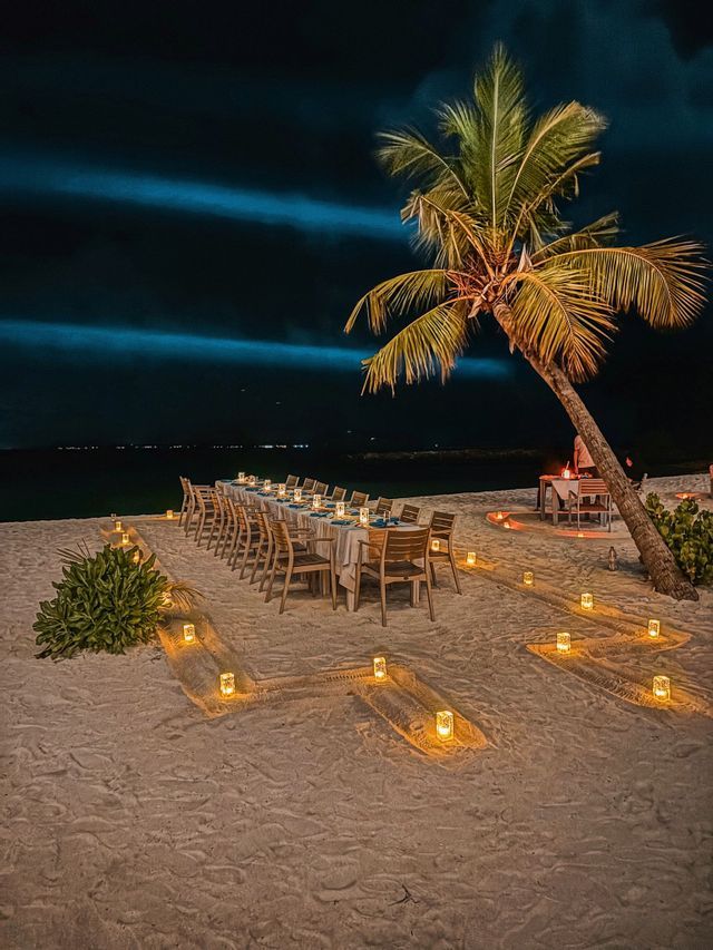 Ein langer Esstisch, nachts auf einem Sandstrand gedeckt und von Kerzen in Laternen beleuchtet, wobei 'WeRoad' im Sand buchstabiert wird.