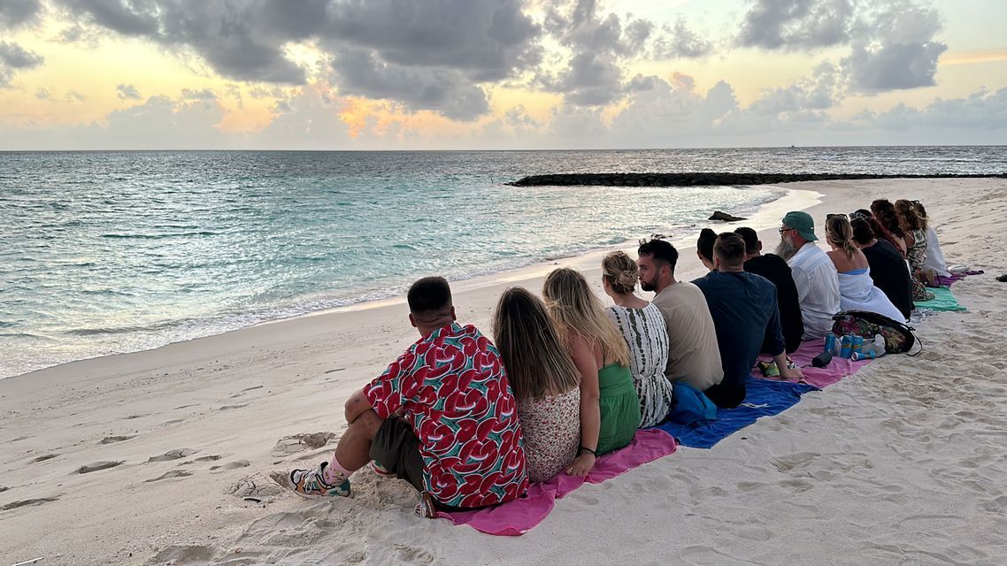 Eine WeRoad-Reisegruppe liegt in einer langen Reihe auf Handtüchern an einem Sandstrand und beobachtet den Sonnenuntergang über dem Meer.