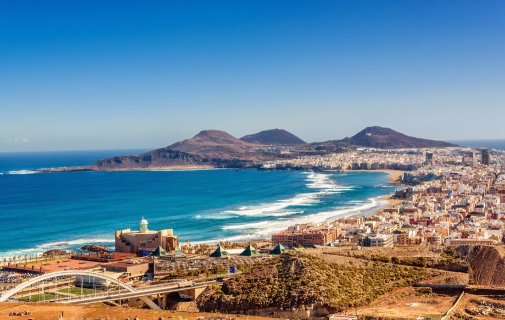 Una vista panoramica di una città costiera lungo una spiaggia sabbiosa, con un oceano turchese e montagne aride sotto un cielo azzurro e limpido.