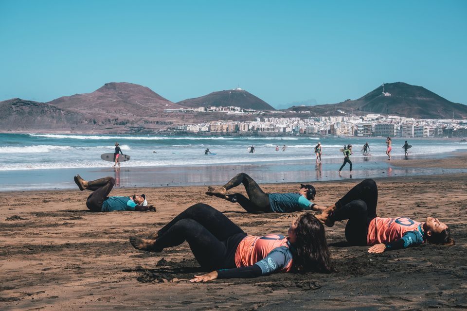 Un viaggio di gruppo WeRoad di persone in muta che fanno stretching su una spiaggia sabbiosa, con surfisti nell'oceano e una città sullo sfondo.