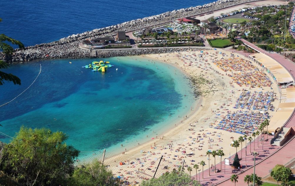 Veduta aerea di una spiaggia sabbiosa affollata a forma di mezzaluna, con persone che prendono il sole sotto ombrelloni colorati e nuotano in acque turchesi.