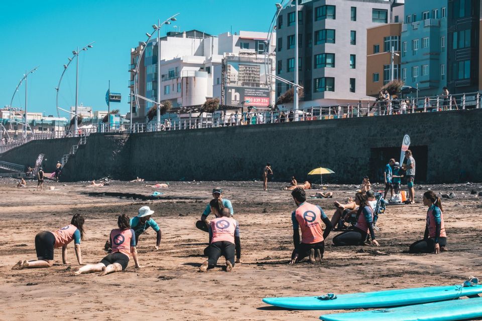 Un viaggio di gruppo WeRoad si prepara su una spiaggia sabbiosa per una lezione di surf, con tavole da surf in primo piano e una città sullo sfondo.