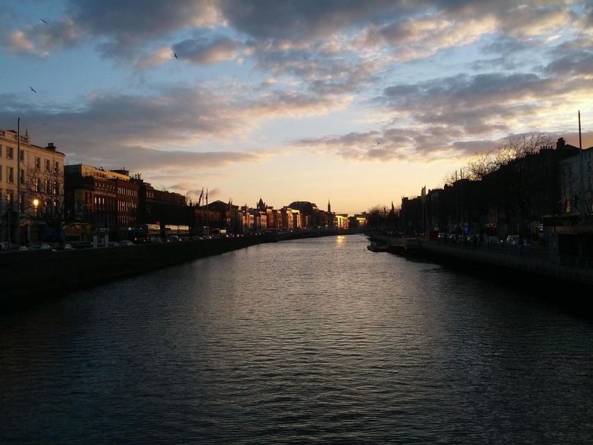Un ampio fiume scorre attraverso una città al tramonto, con edifici che costeggiano le sponde sotto un cielo nuvoloso.